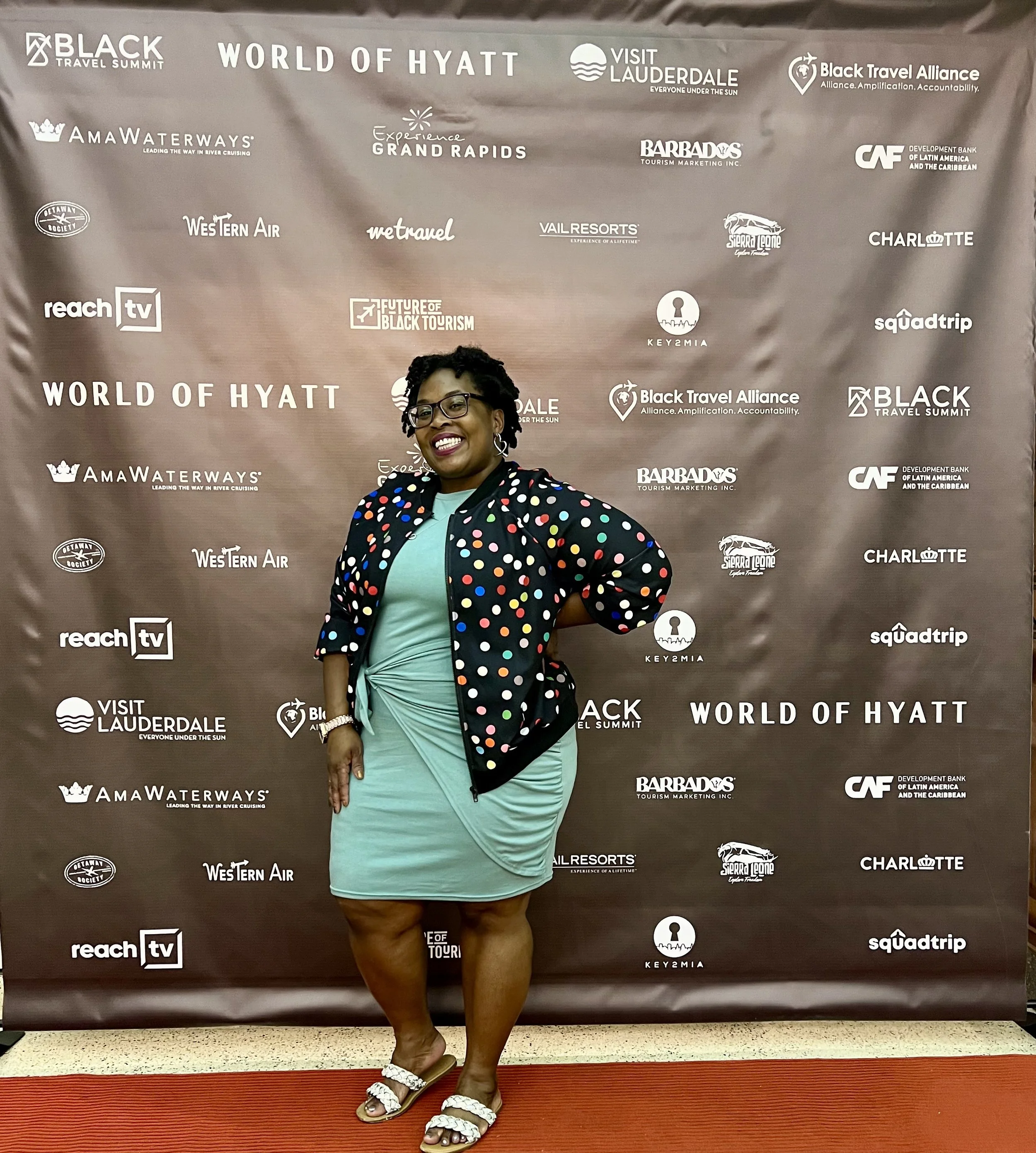 Leslie in a teal dress and polka dot jacket standing in front of the backdrop for the Black Travel Film Festival